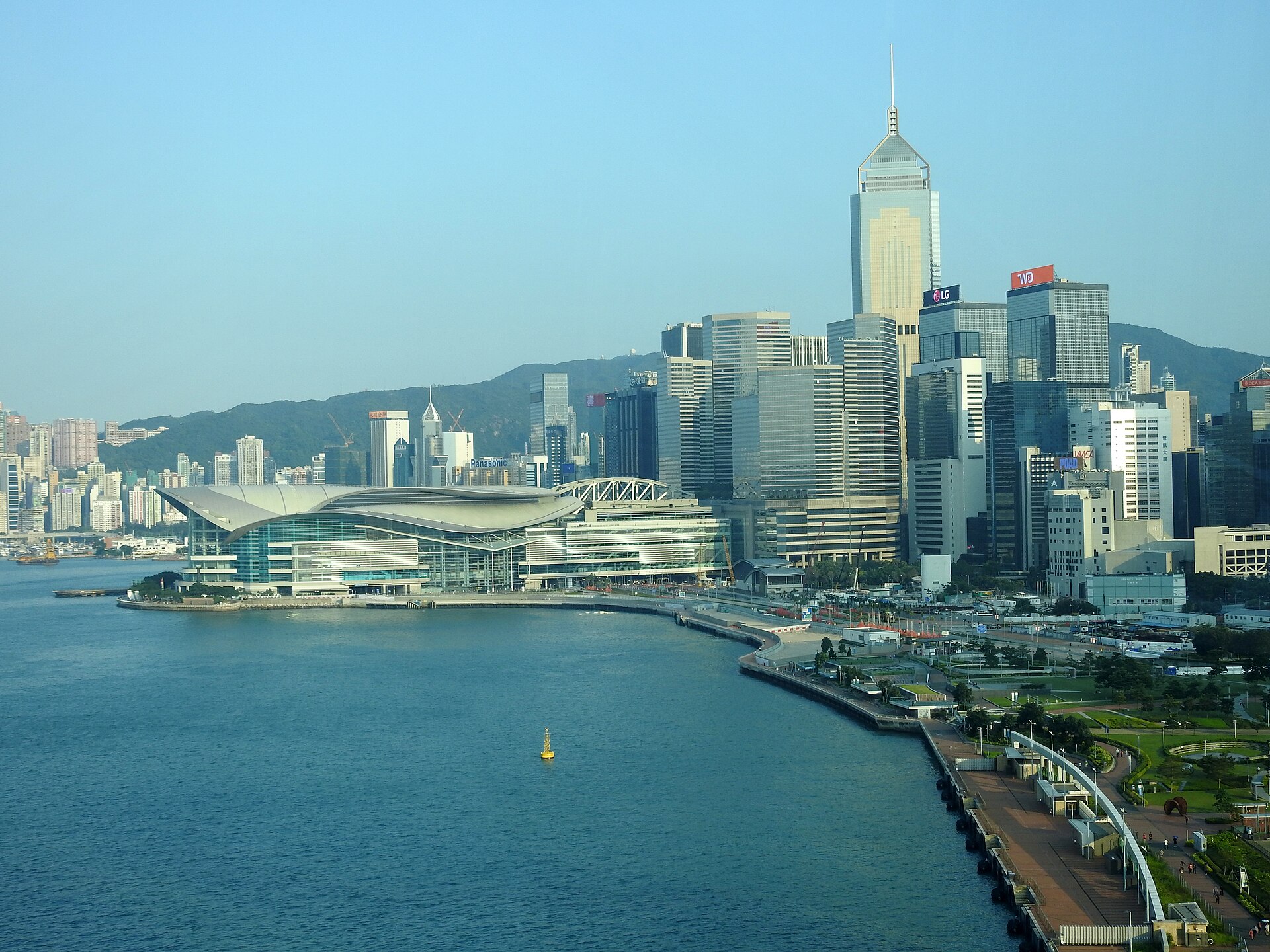 Aerial view of the curved-roof Hong Kong Convention and Exhibition Centre on the harbor waterfront, with office towers and green hills behind.