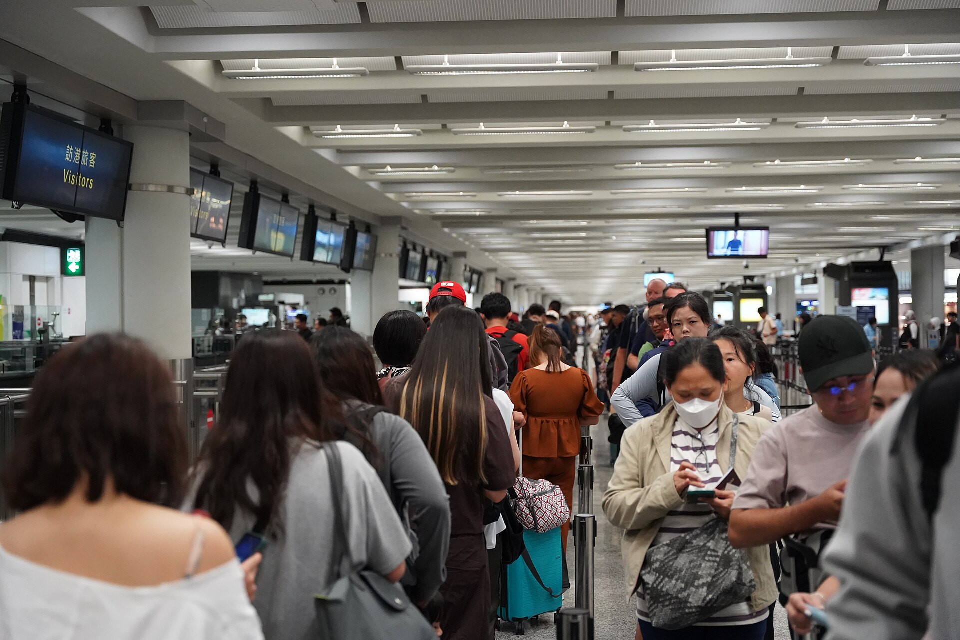 Long lines of travelers with bags and phones waiting in the immigration hall at Hong Kong International Airport, under a sign reading Visitors