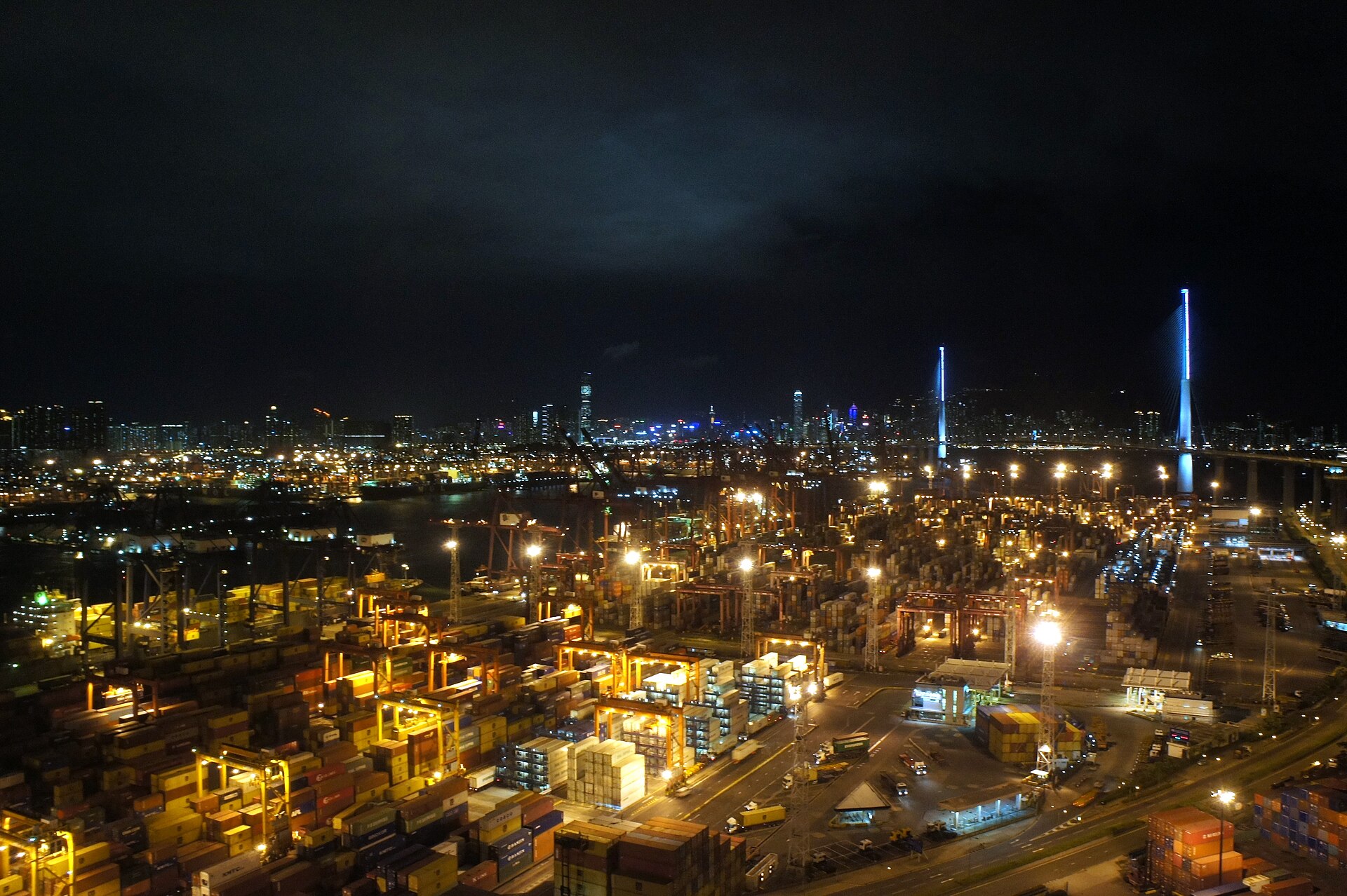 Night view over a floodlit container yard with stacked shipping containers and gantry cranes, a city skyline across the water and the tall lit towers of a cable-stayed bridge at right